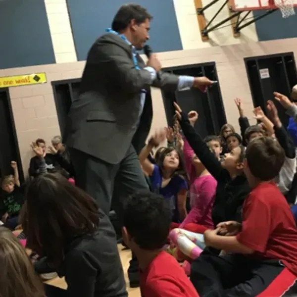 A man in a suit is speaking into a microphone in front of a crowd of children