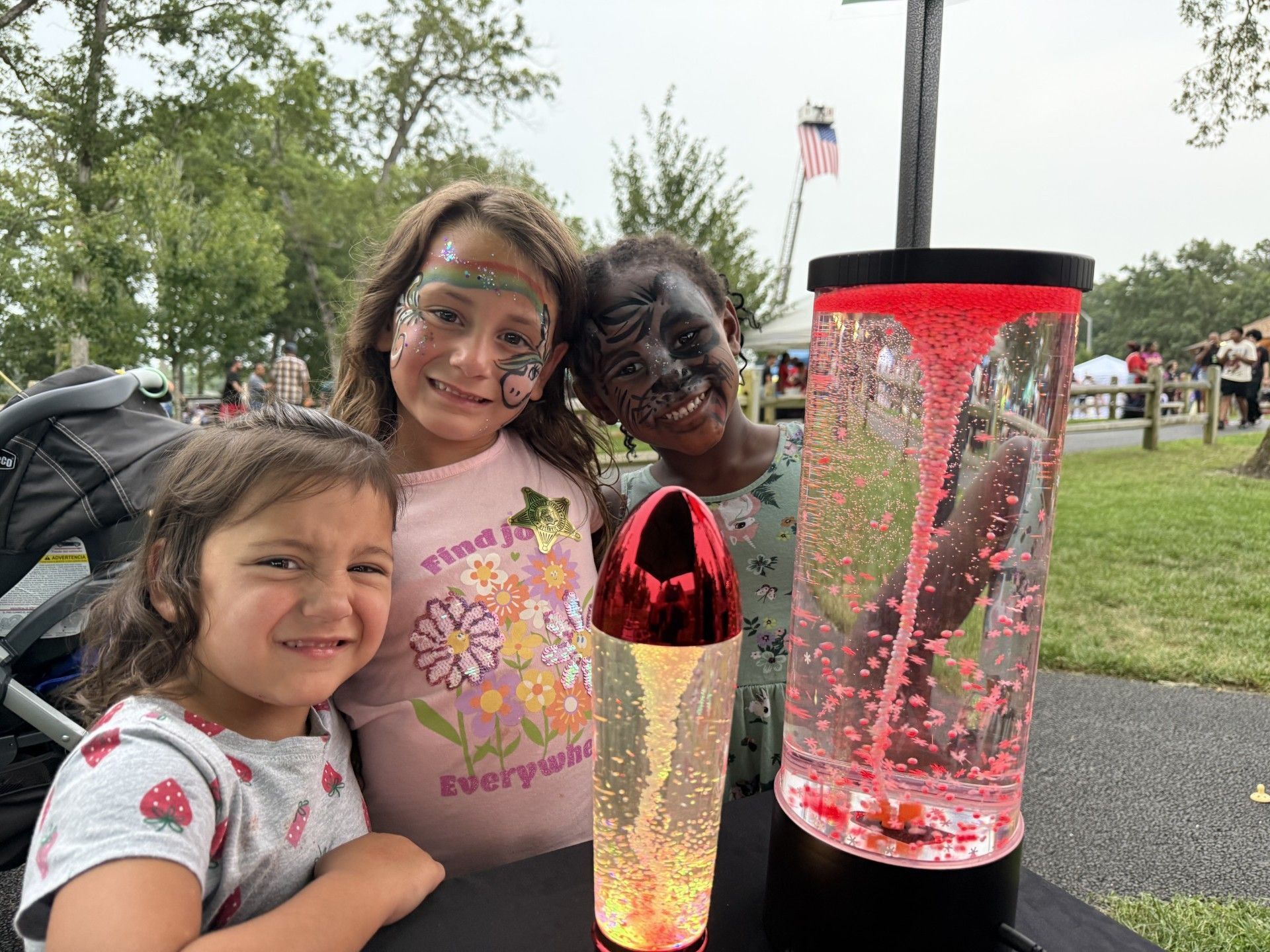 Three smiling children with face paint pose near a glowing vortex tube at an outdoor event.