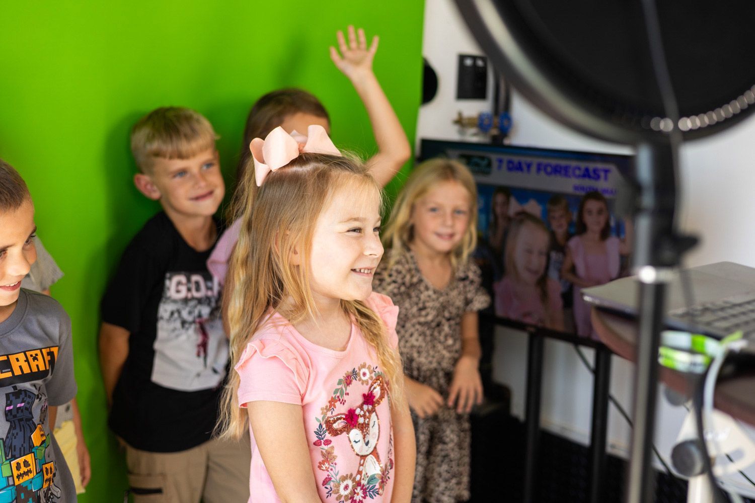 Children in front of a green screen, some waving and smiling. Weather forecast on a monitor behind them.