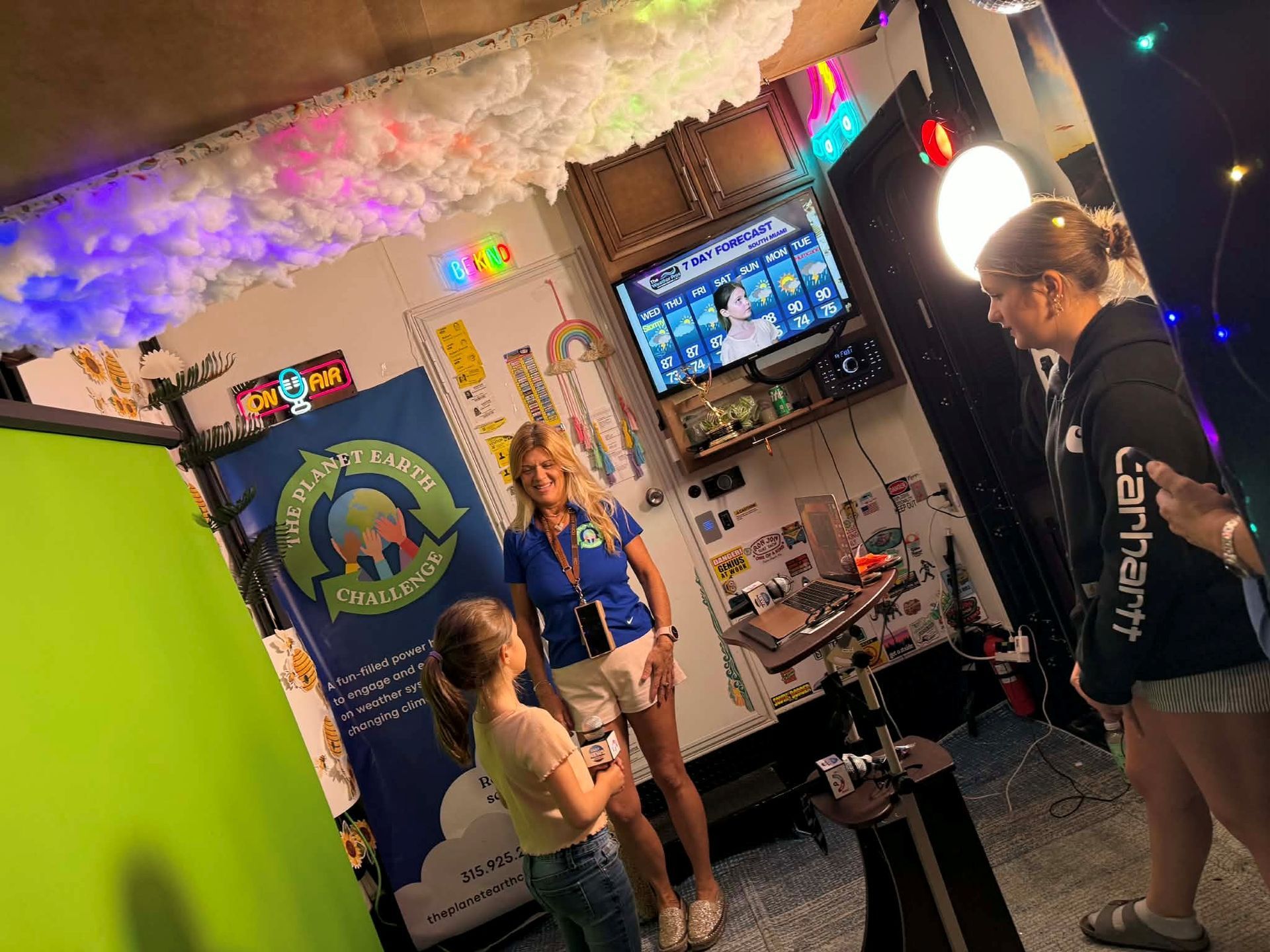 A girl and two women on a set with a green screen, camera, and TV. The set has a rainbow theme. 