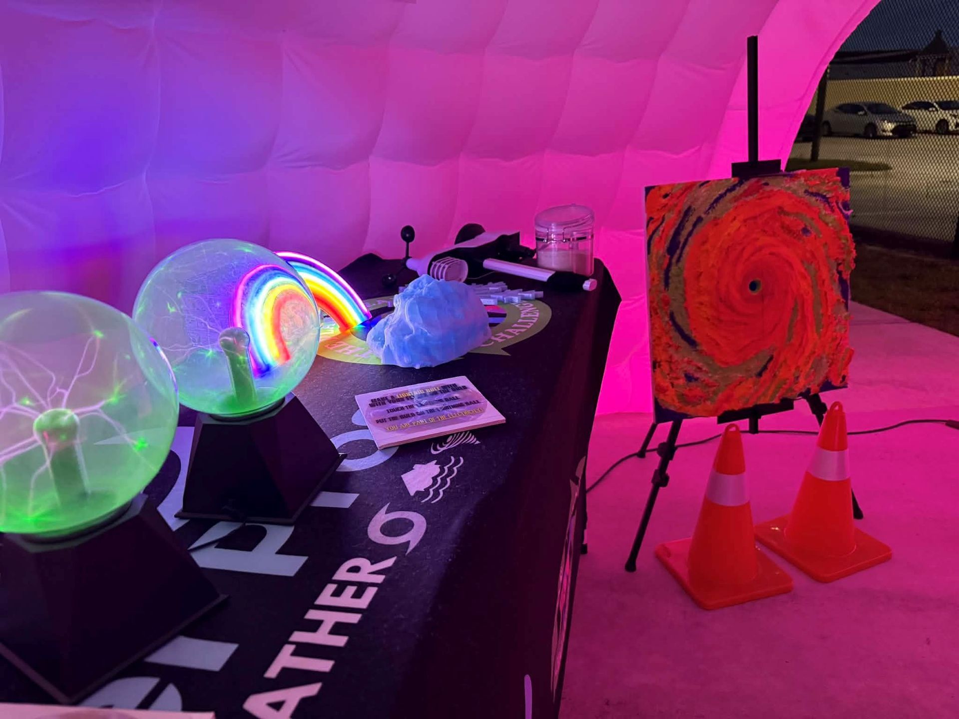 A weather-themed display with plasma balls, a painted spiral, and safety cones, lit by pink and purple lights. 
