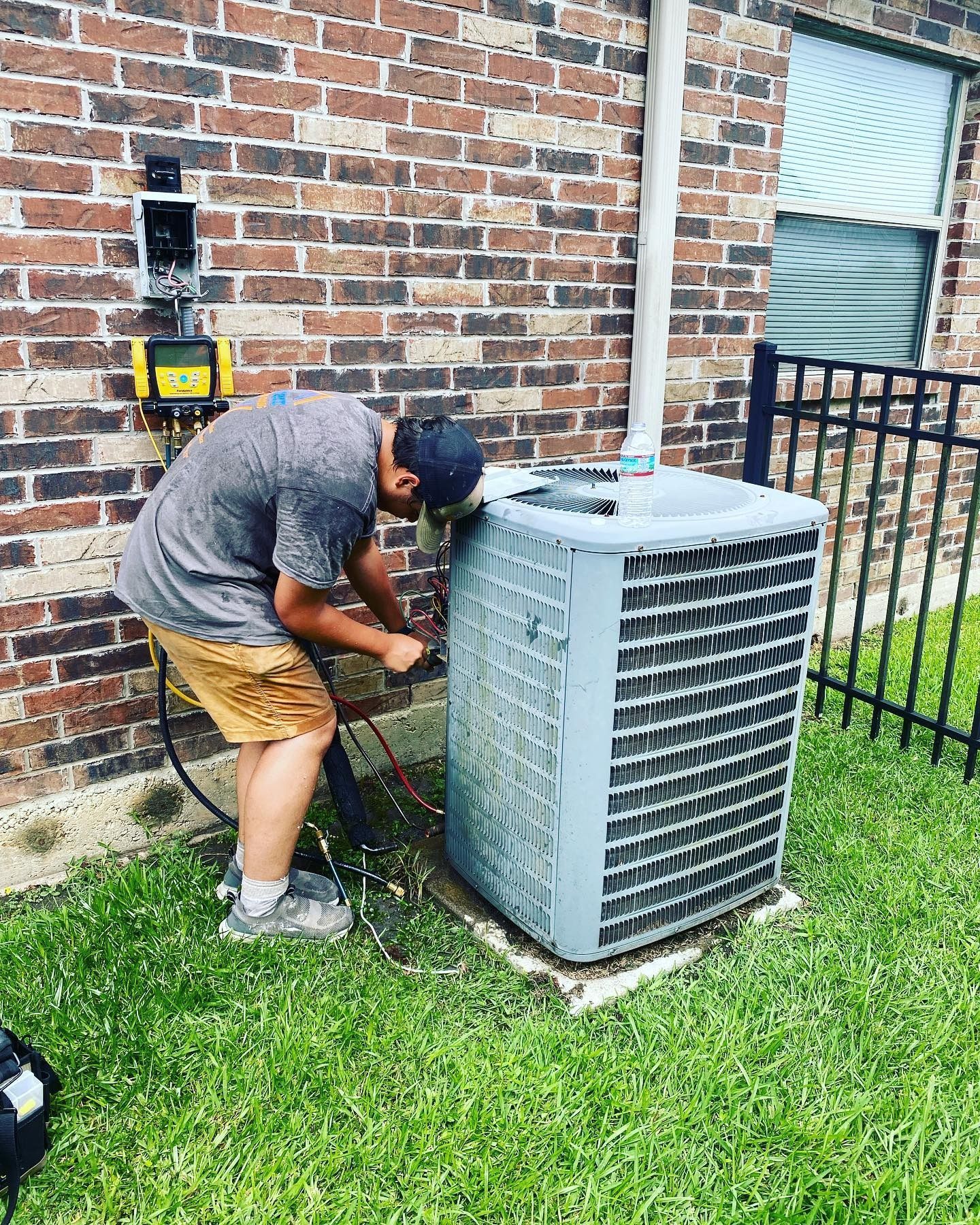 A technician works on an outdoor HVAC unit mounted on a concrete pad next to a brick wall.
