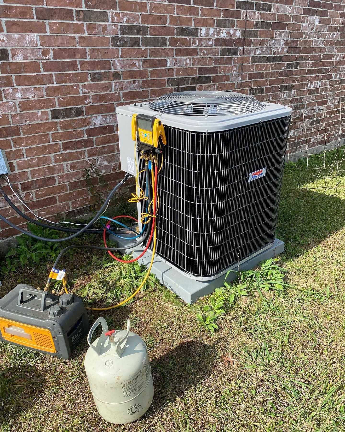 An HVAC technician’s gauge set and recovery machine are connected to an outdoor air conditioner unit for maintenance.