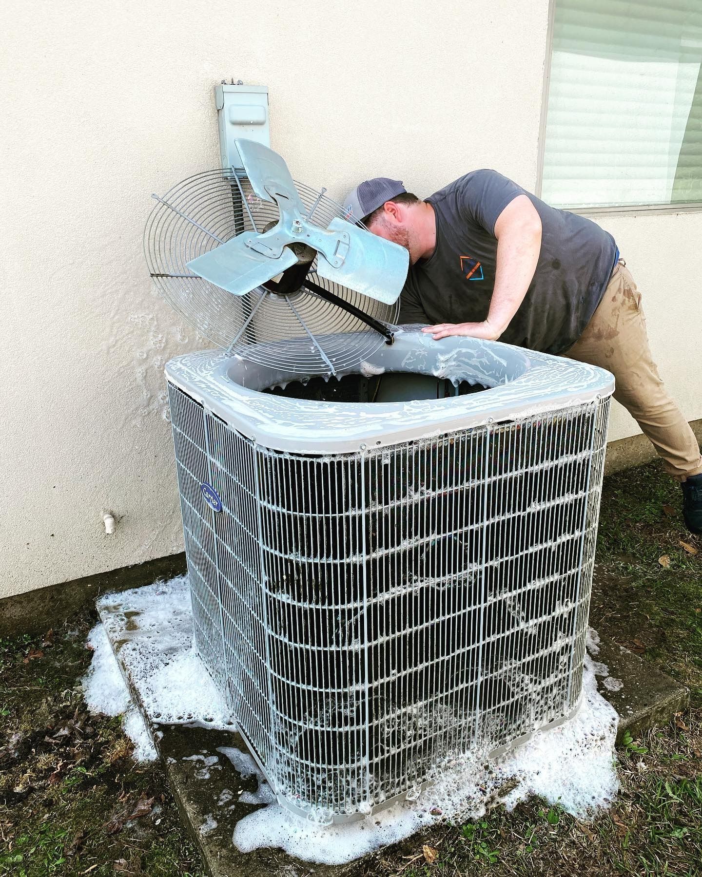 A person in a gray shirt leans over an outdoor air conditioning unit covered in thick, white cleaning foam.