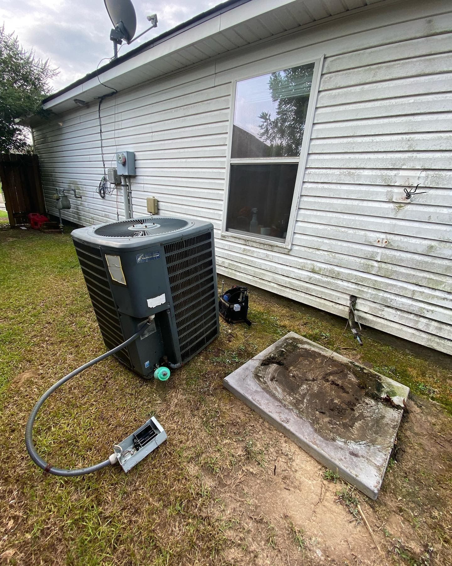 An outdoor HVAC condenser unit sits on the grass next to a house, with an electrical box disconnected and lying on the ground.