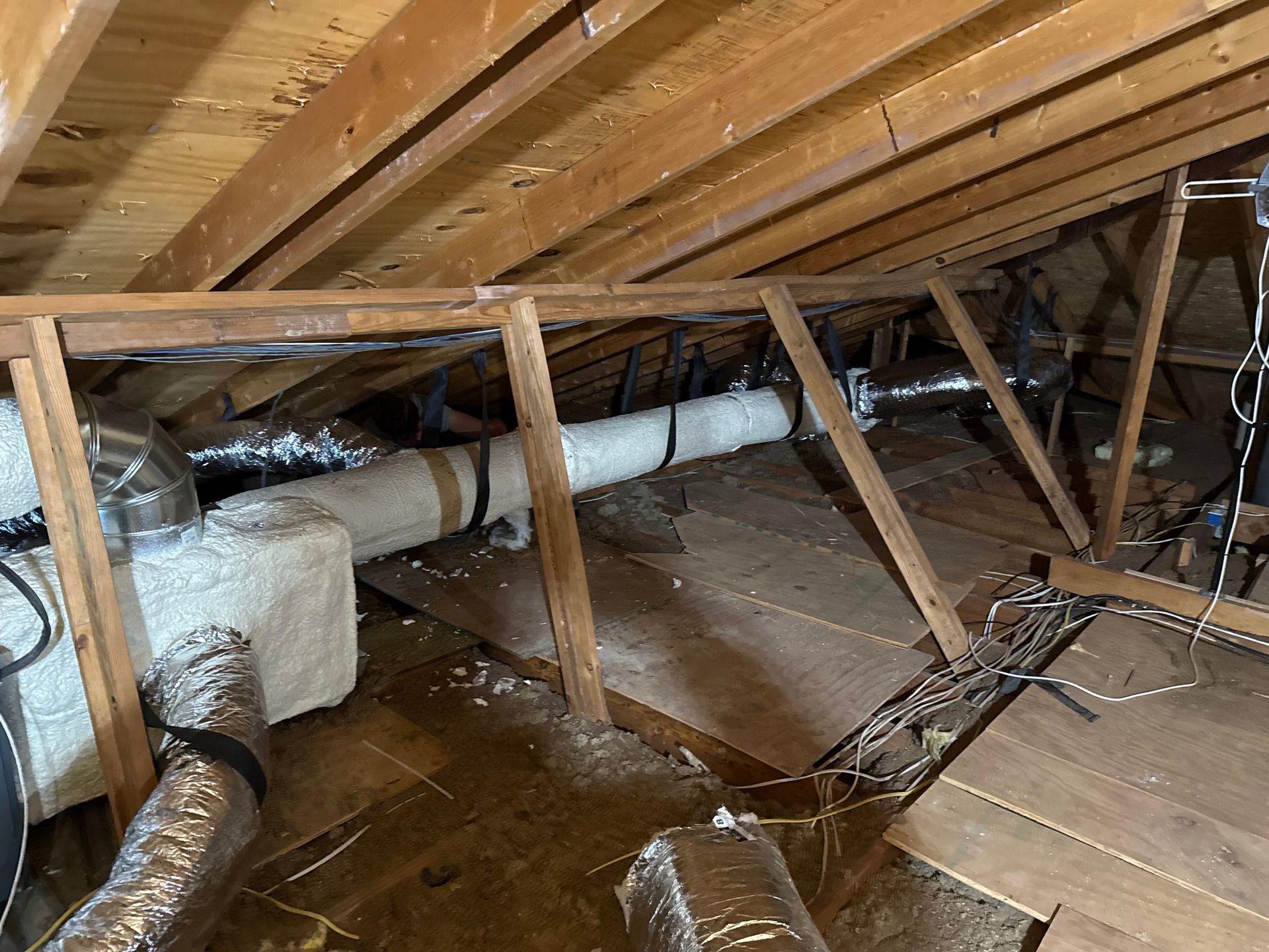 An unfinished attic with wooden rafters, insulated HVAC ductwork, and electrical wiring on the floorboards.