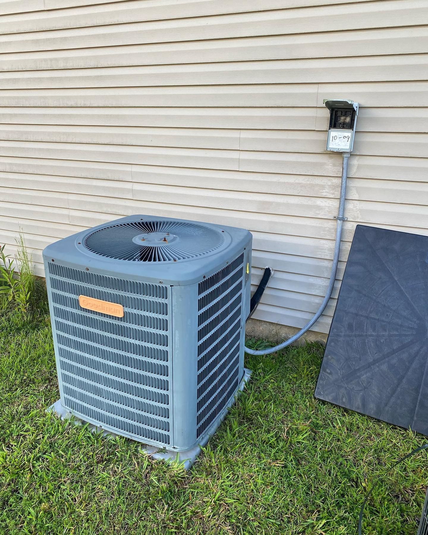 A gray air conditioning unit sitting on grass next to a house siding with a connected electrical disconnect box.