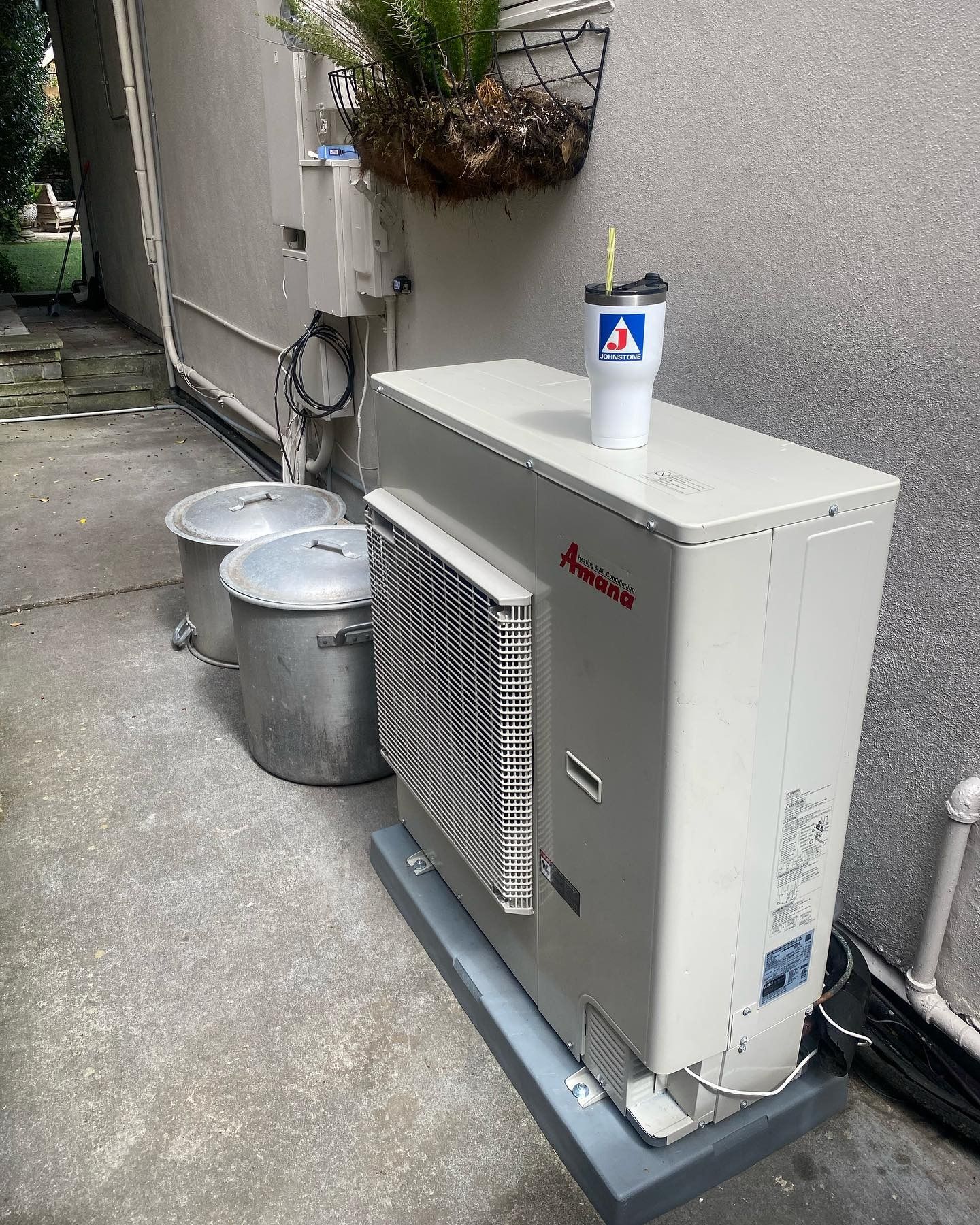 An outdoor beige HVAC unit sits on a concrete pad next to two metal pots, with a white tumbler cup on top of the unit.
