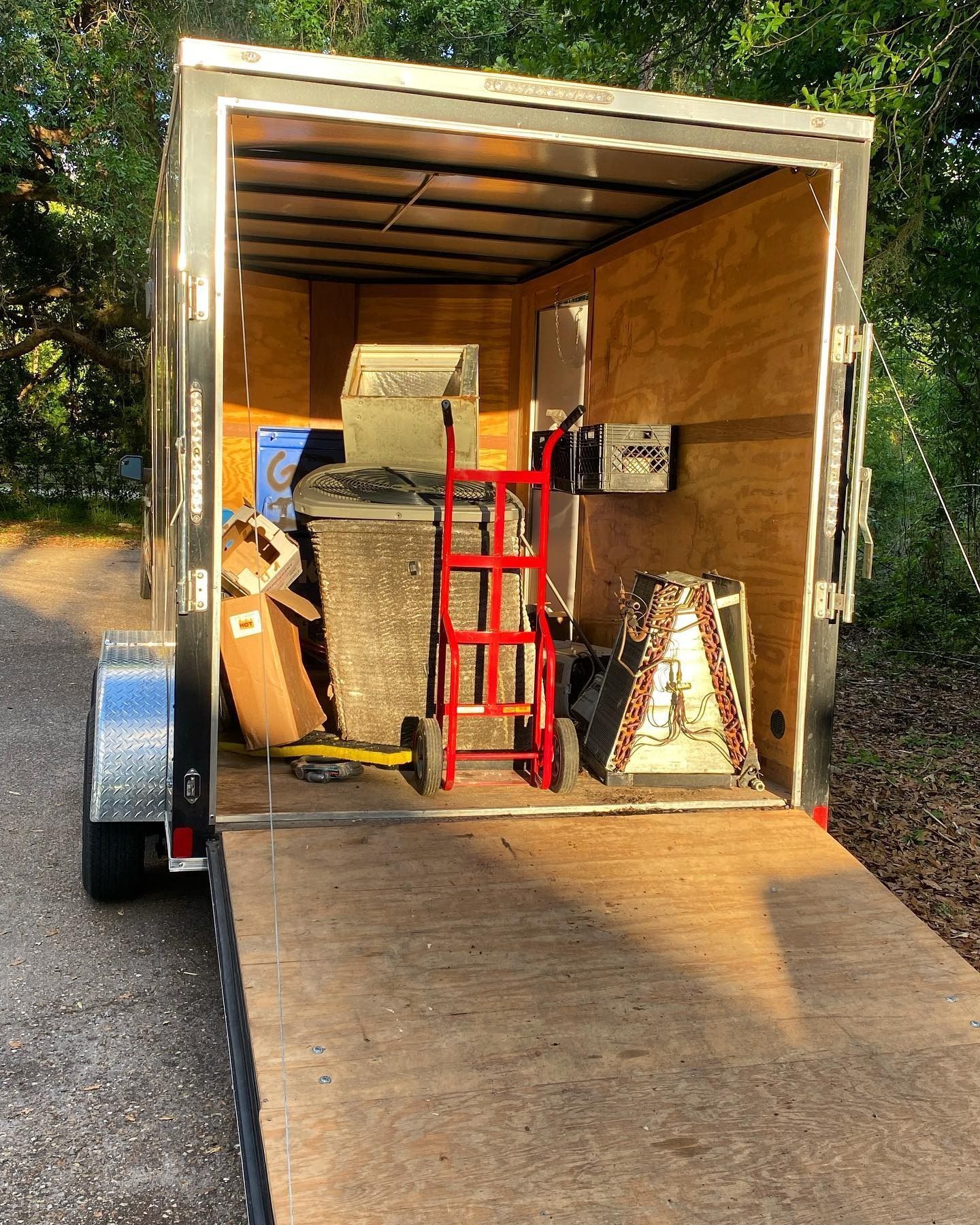 A view into an open cargo trailer containing a red hand truck and various household items parked on a gravel path.