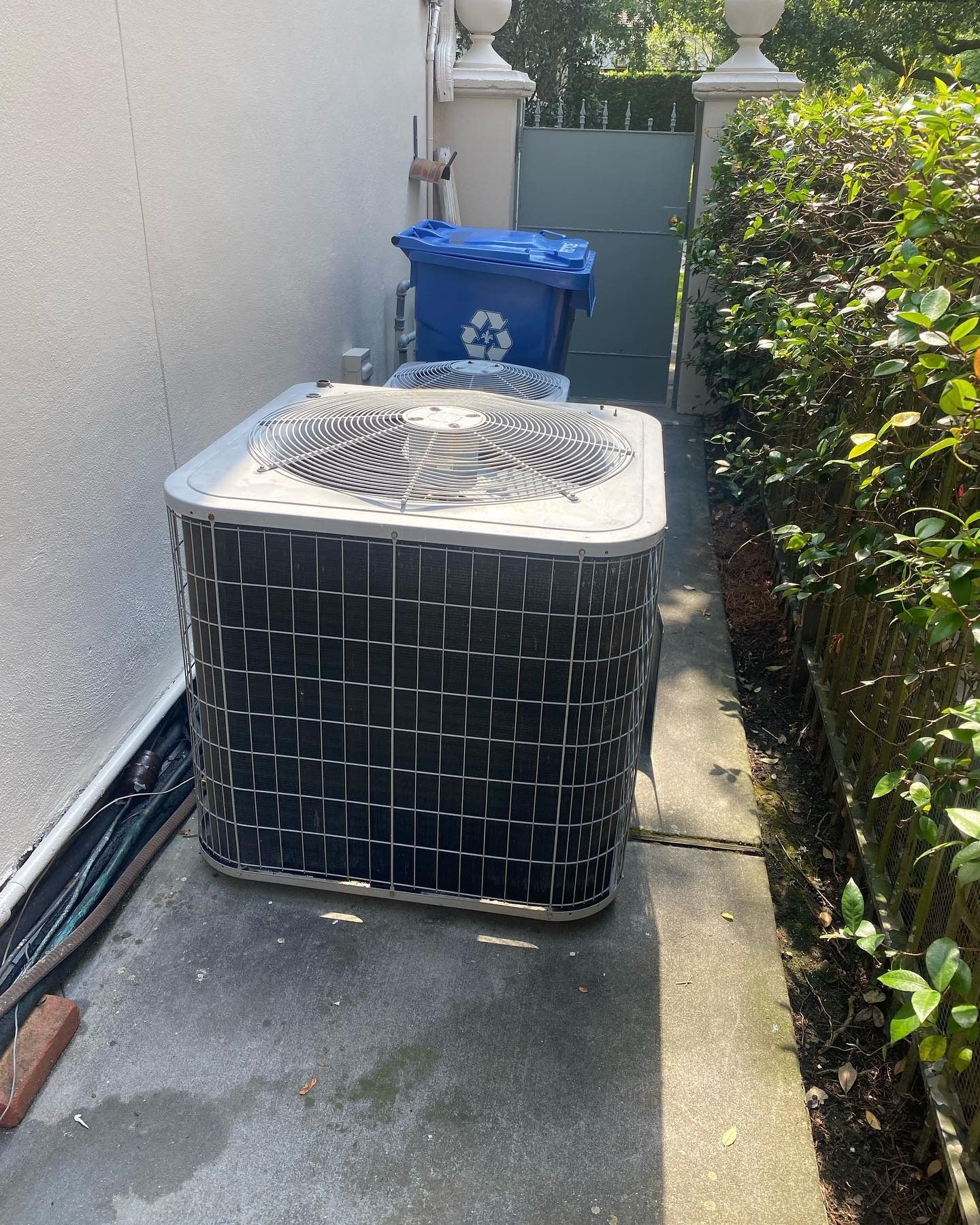 A square outdoor air conditioning unit sits on a concrete path beside a wall, with a recycling bin visible in the back.