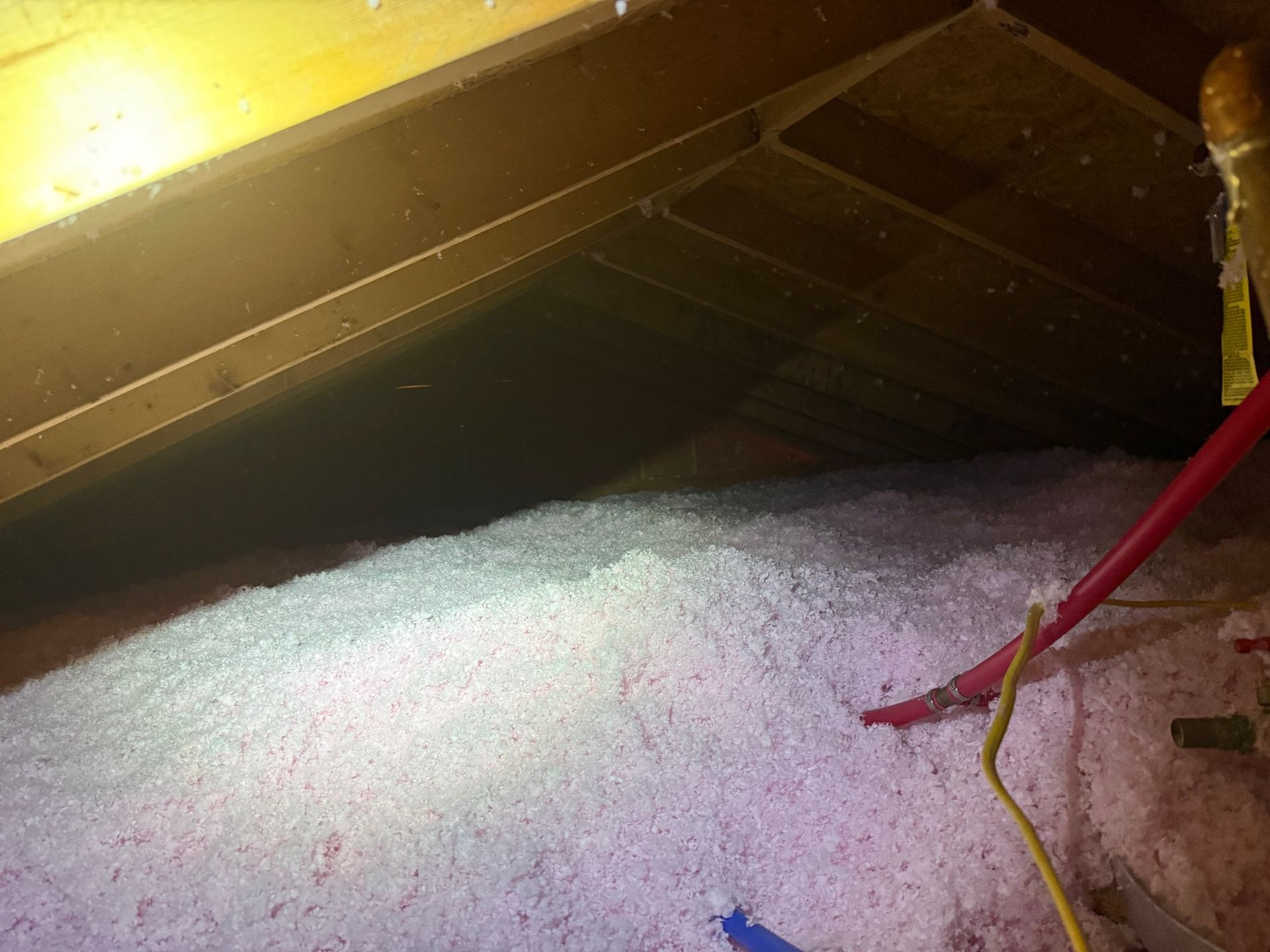 Attic space with pink blown-in insulation covering the floor, showing roof rafters and electrical cables.