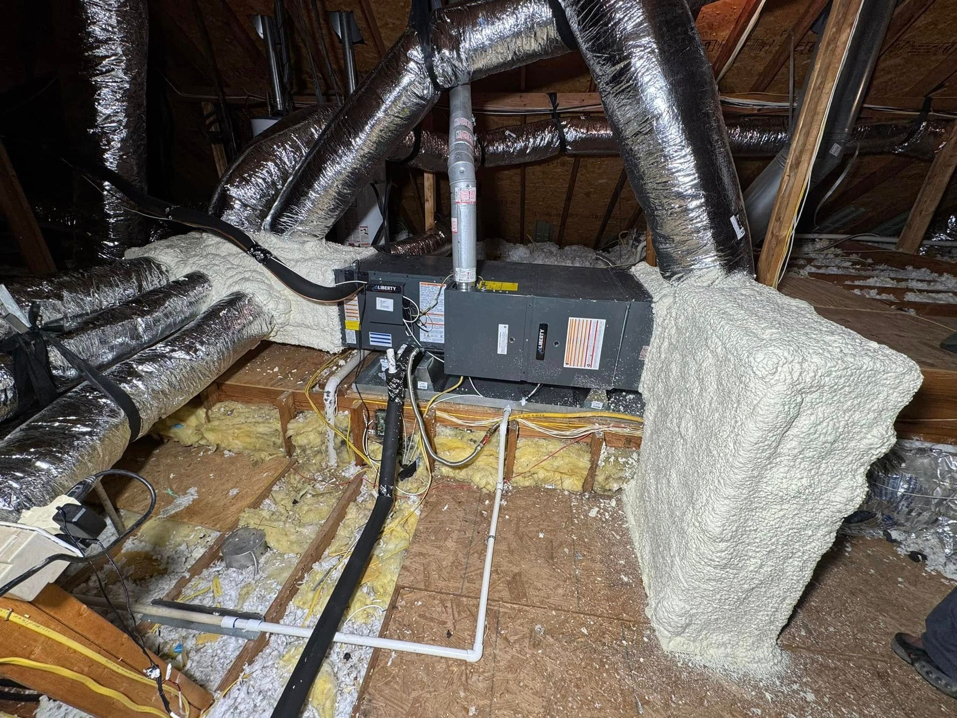 An HVAC air handler unit installed in an attic, surrounded by silver ductwork and thick fiberglass insulation.