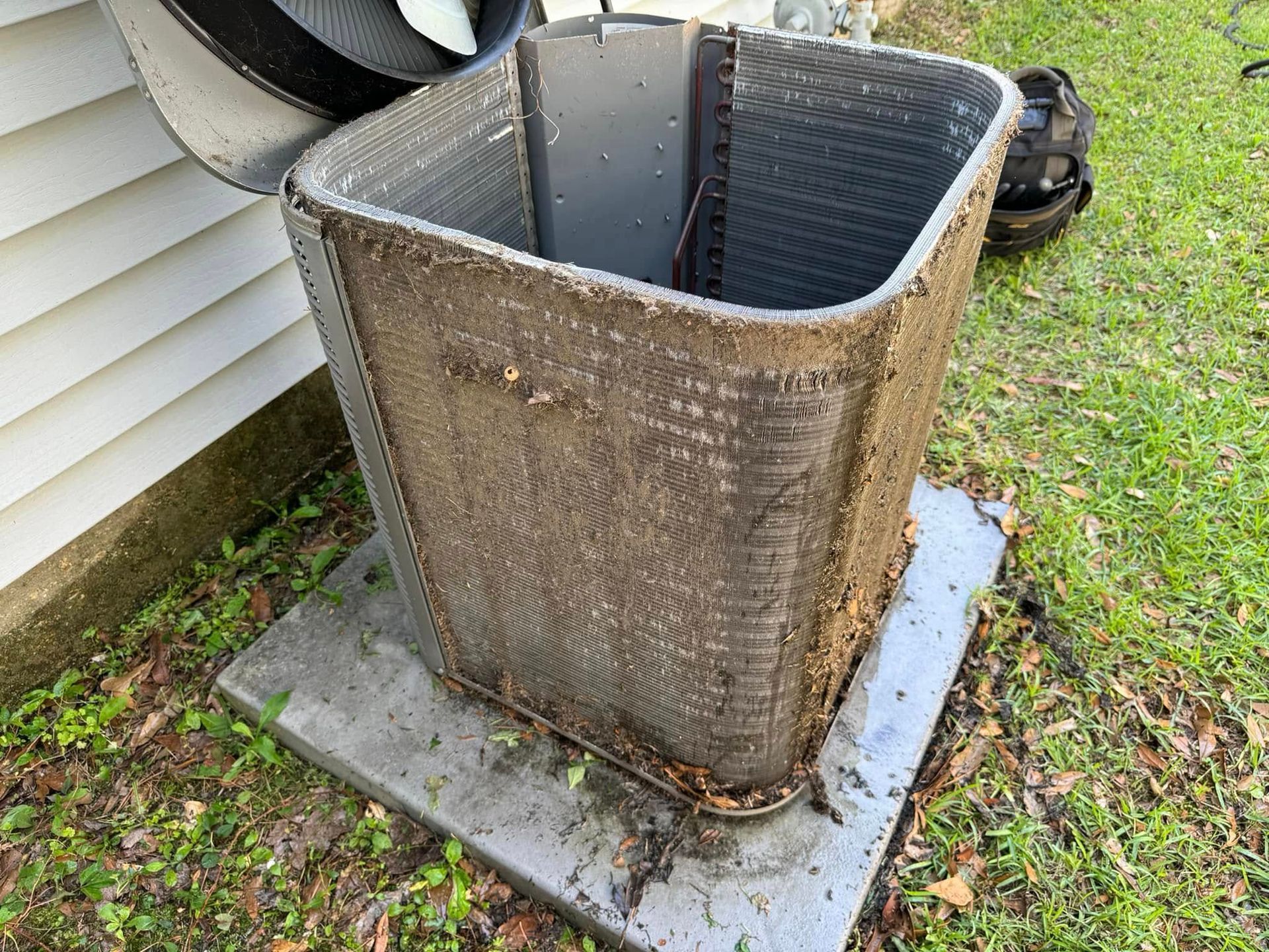 A dirty, clogged air conditioning condenser coil unit sitting on a concrete slab outdoors next to a house.