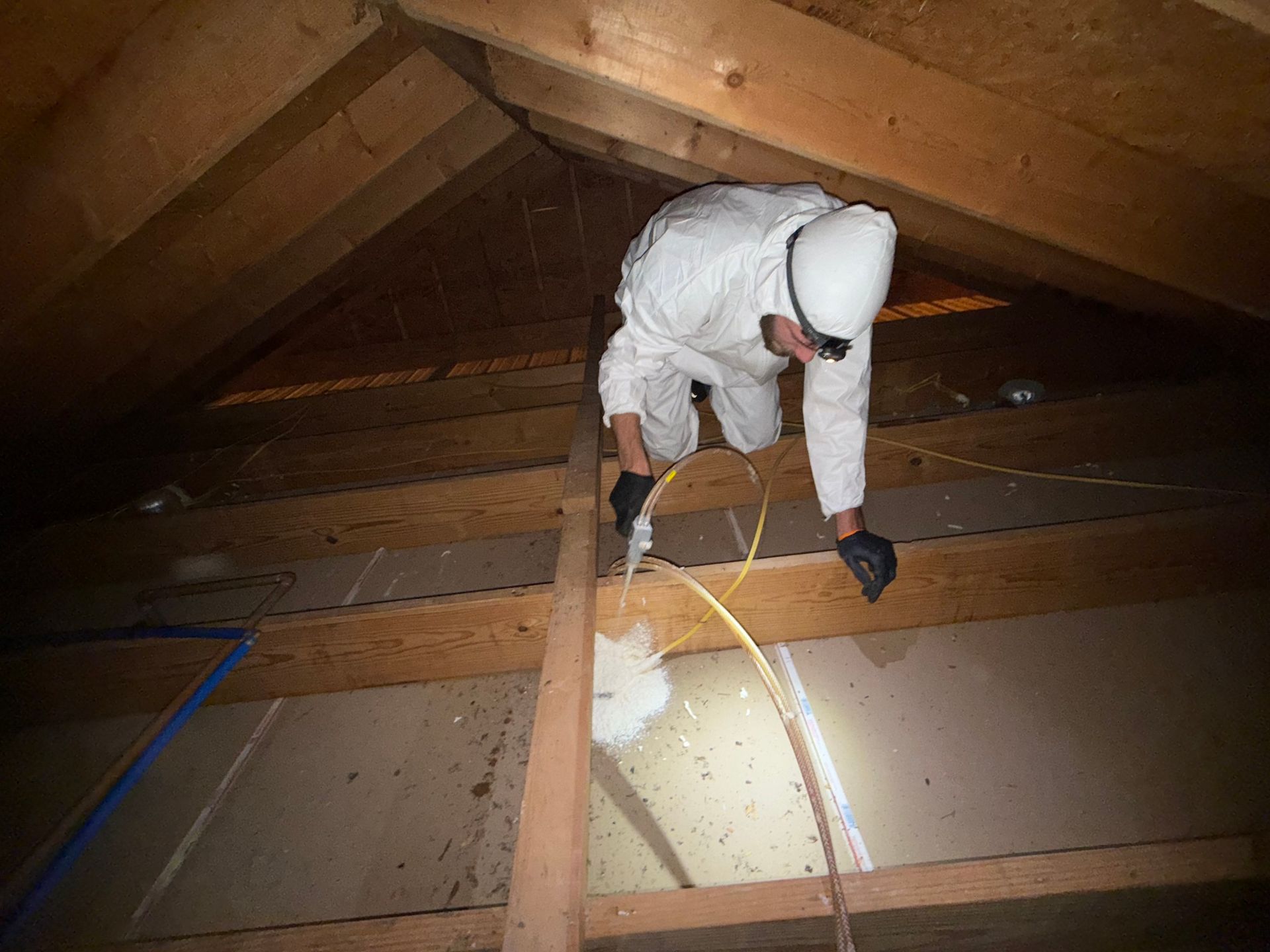 A person in protective gear applies foam sealant to a joint in a wooden attic frame.