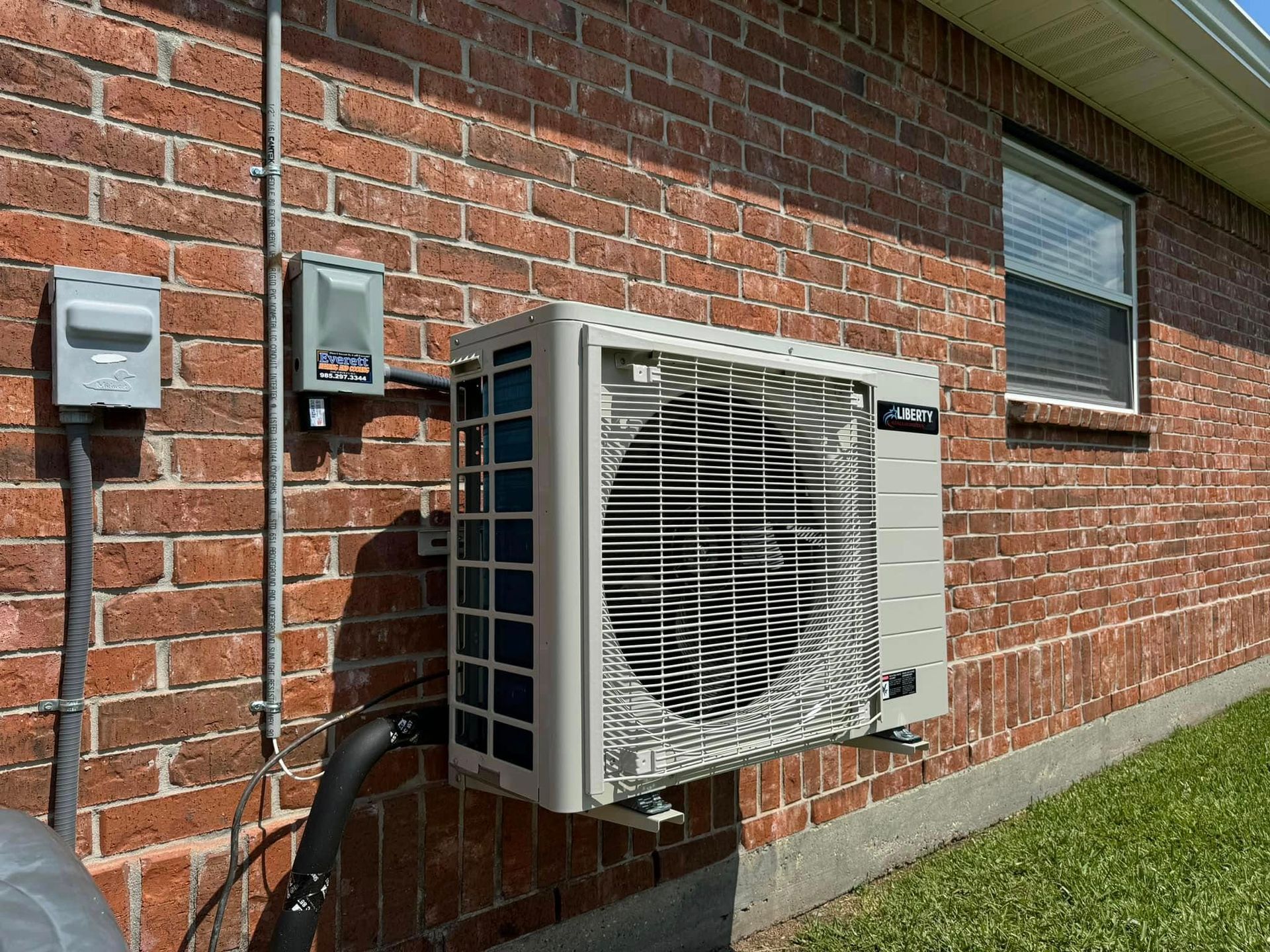 A tan outdoor HVAC condenser unit mounted on a brick wall next to an electrical disconnect box and conduit.