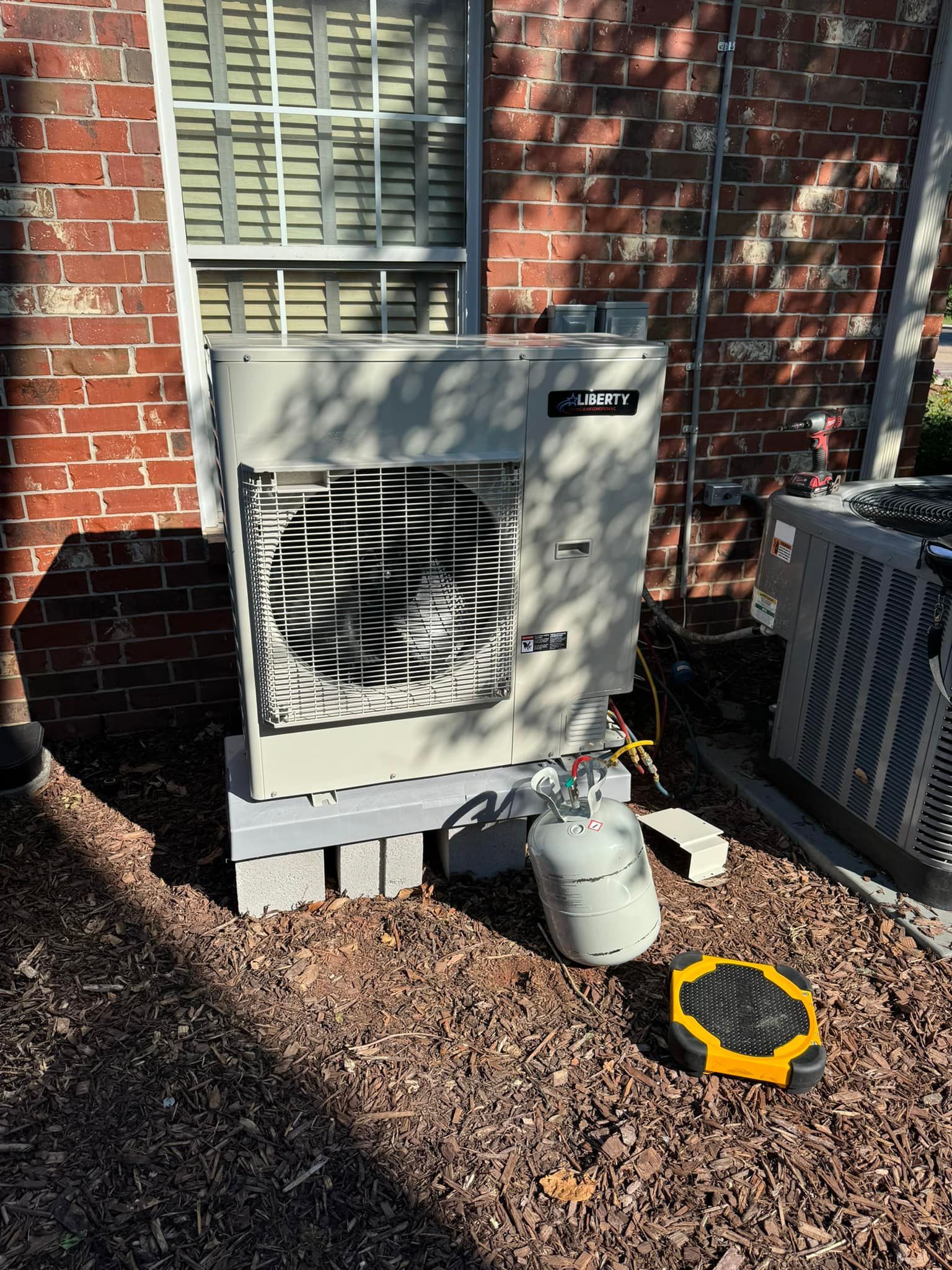 An outdoor HVAC unit sitting on concrete blocks next to a brick wall, with a small refrigerant cylinder nearby.