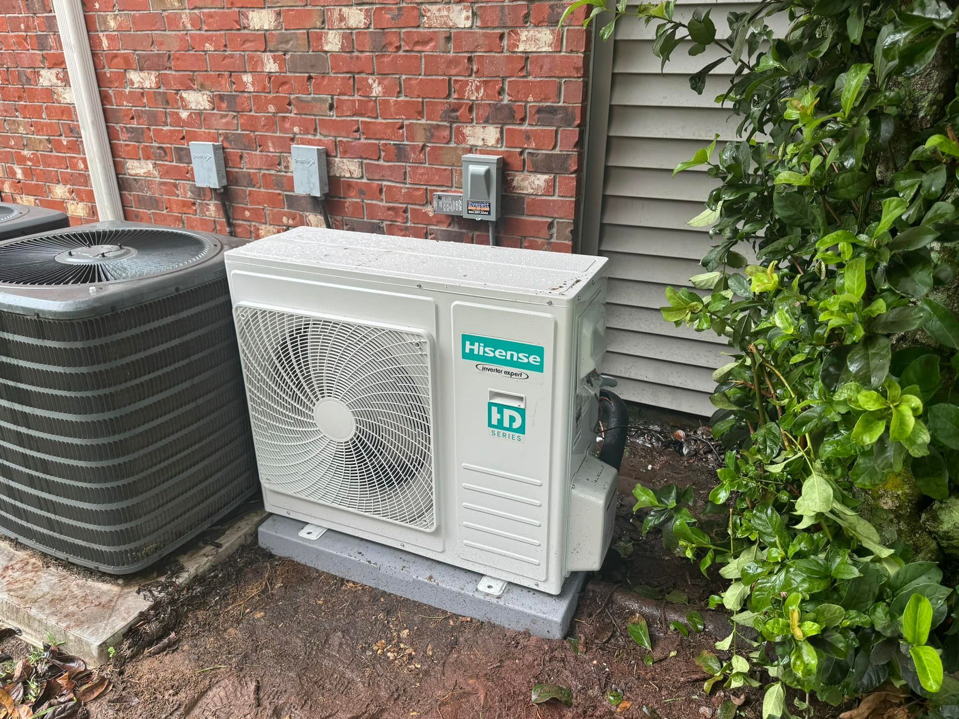 A white outdoor HVAC heat pump unit sits on a concrete pad against a brick wall, next to a larger dark grey air conditioner.