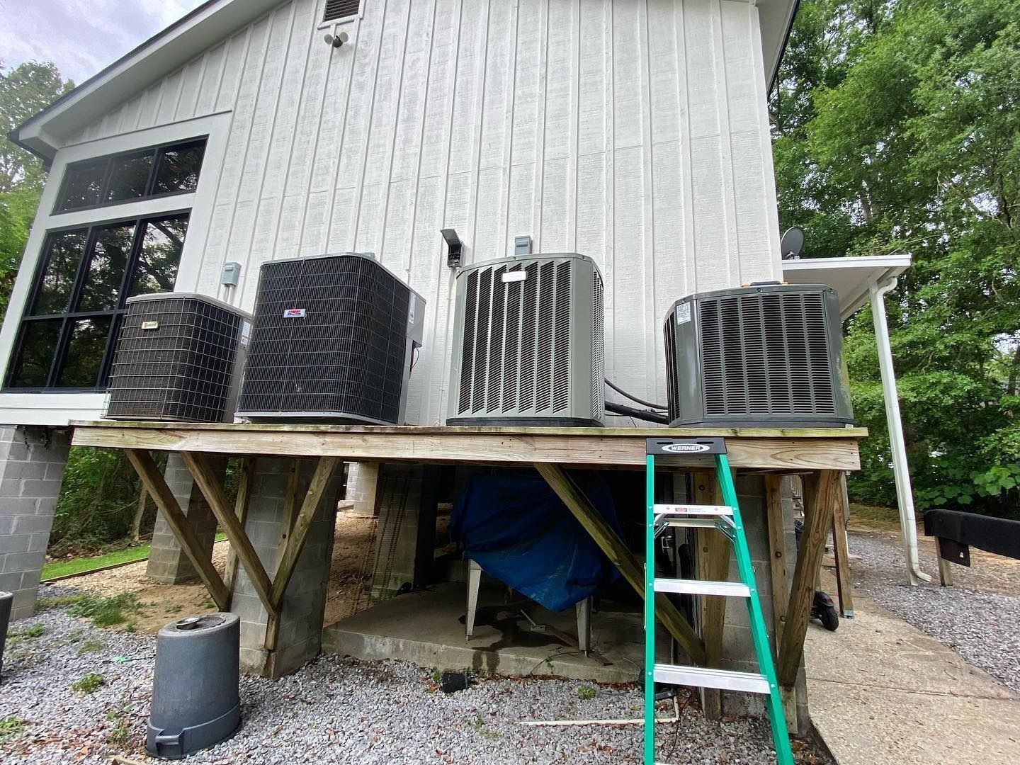 Four air conditioning condenser units mounted on an elevated wooden platform next to a white house.
