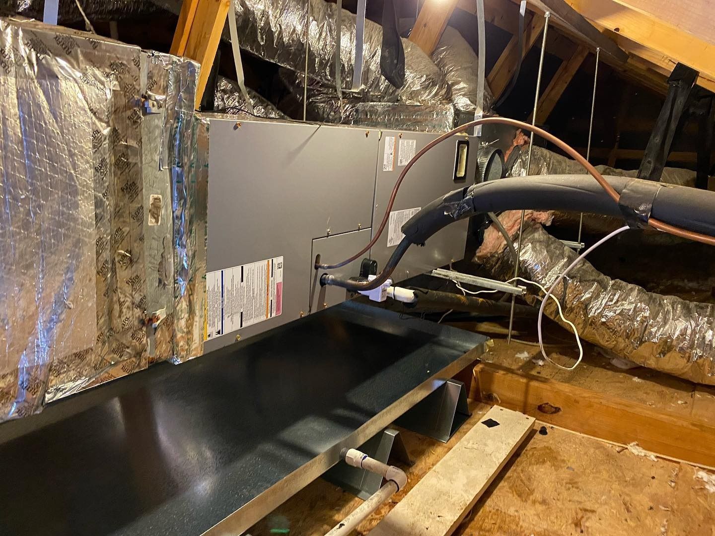 An HVAC air handler unit installed in an attic, resting over a metal emergency drain pan with connected ductwork and piping.