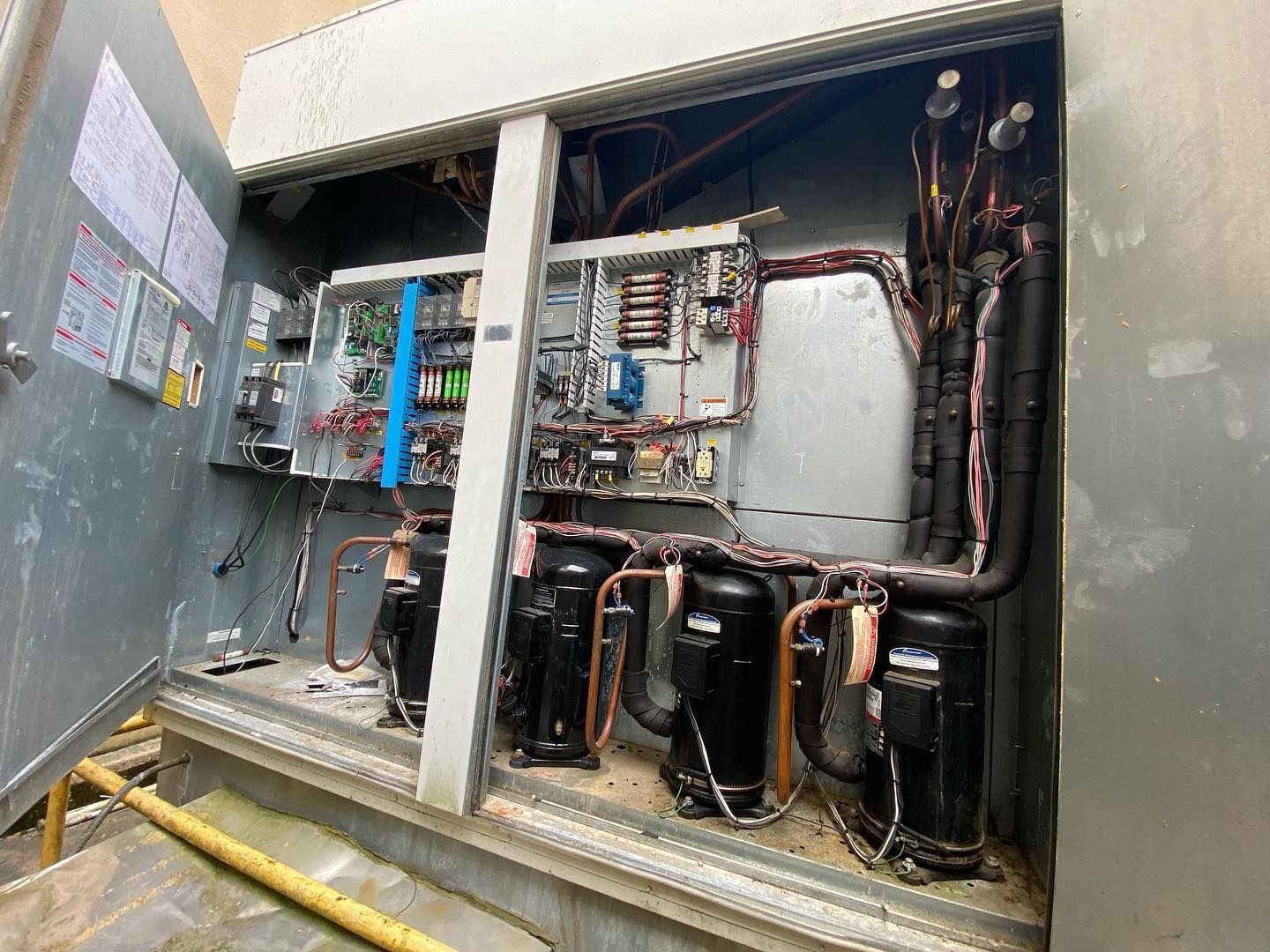 The open electrical compartment of an HVAC rooftop unit shows circuit boards, wiring, and four black compressors.