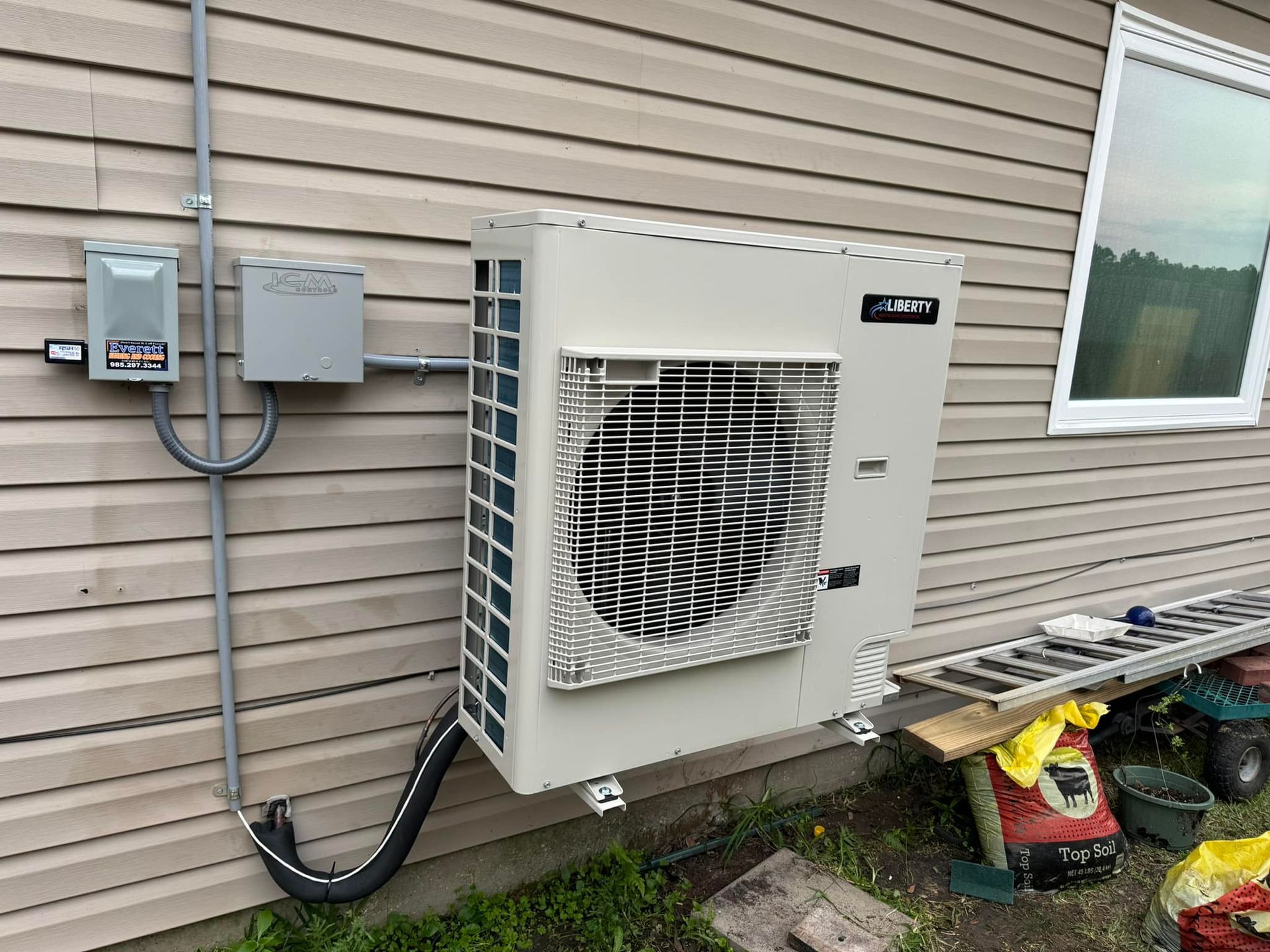 An outdoor mini-split heat pump unit mounted on the side of a house next to electrical boxes and a window.