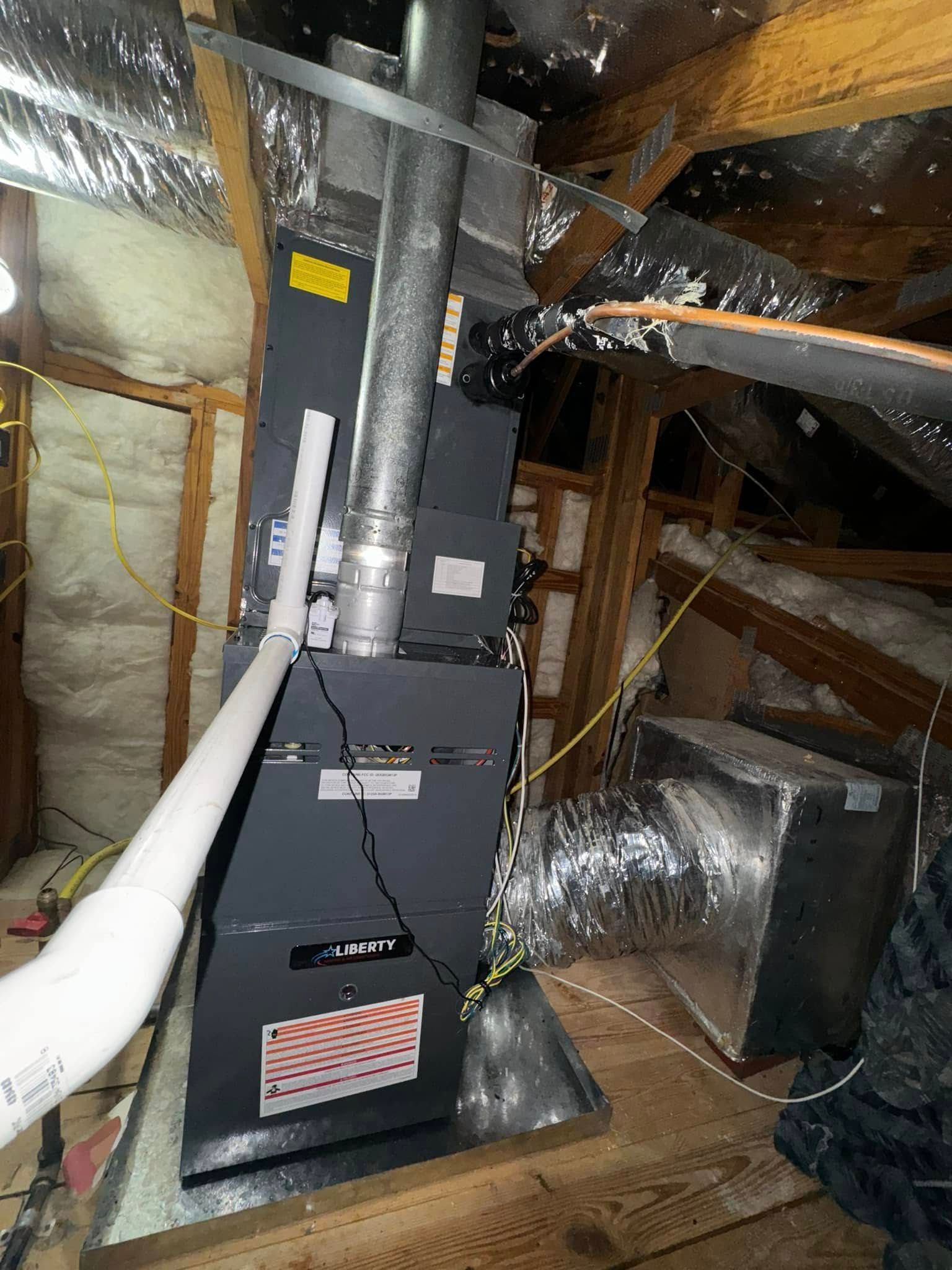 A grey furnace unit with a metal exhaust pipe and white PVC drain line installed in an attic space with insulation.