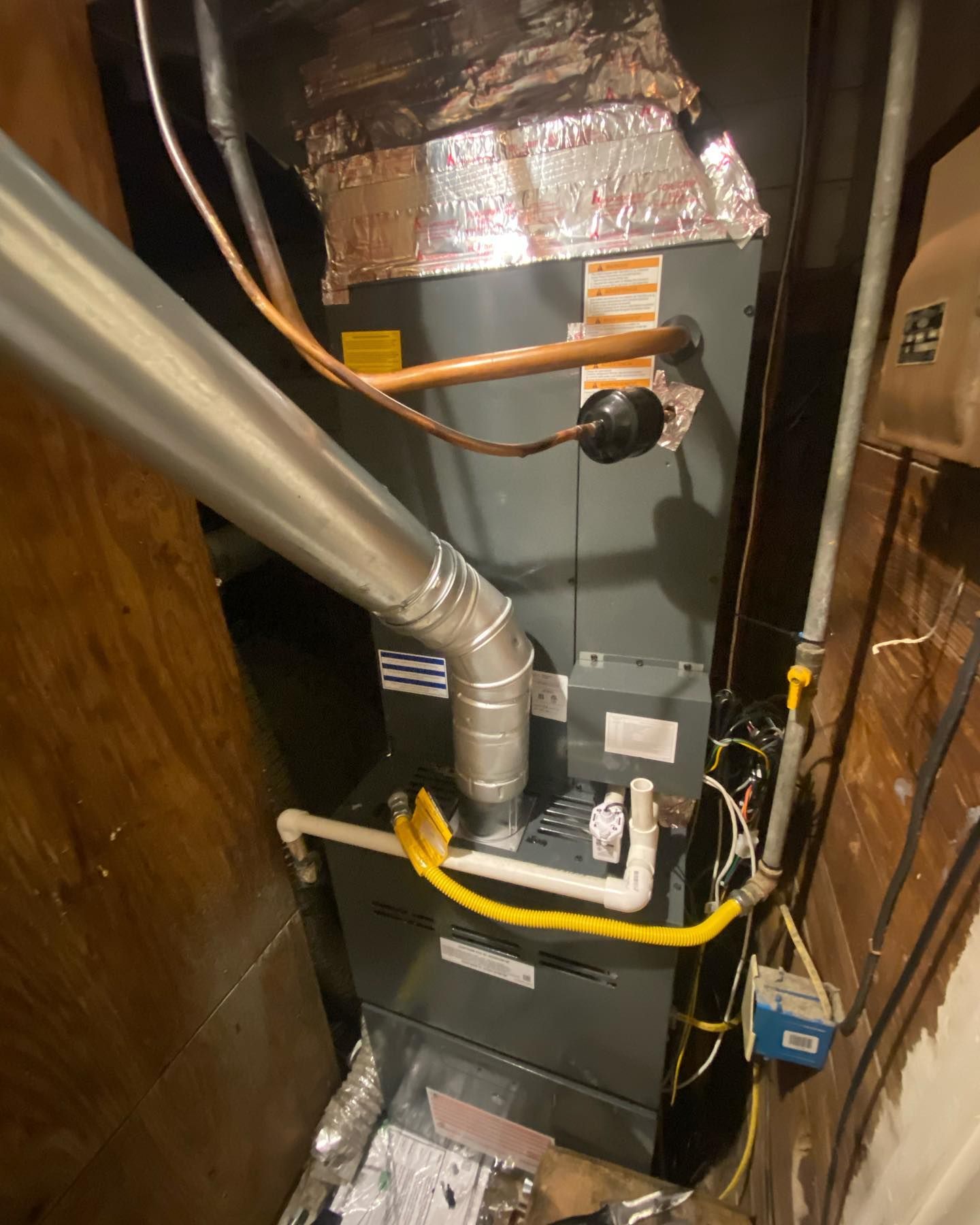 A gray furnace unit in a utility closet with a metal exhaust pipe, copper lines, and white PVC plumbing.