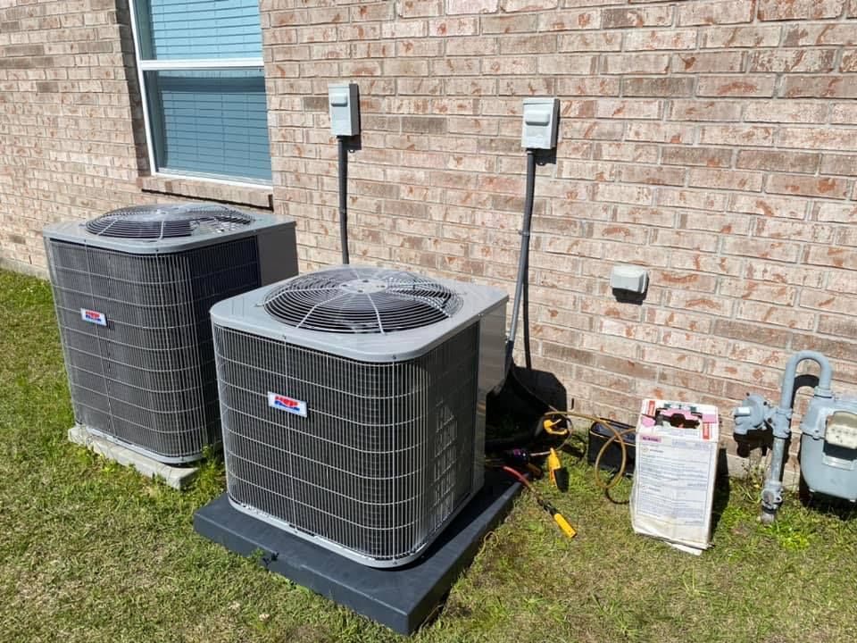 Two rectangular HVAC units sit on a grassy lawn against a brick wall, with power boxes and a gas meter nearby.