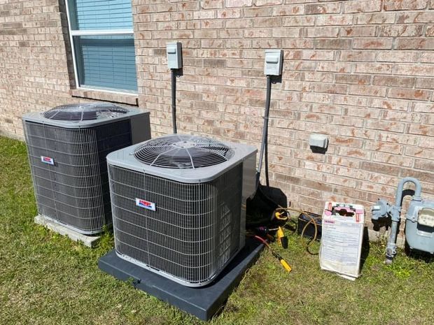 Two HVAC condenser units sit on a lawn next to a brick house wall, with electrical disconnect boxes and a gas meter.