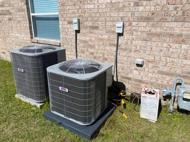 Two HVAC condenser units sit on a lawn next to a brick house wall, with electrical disconnect boxes and a gas meter.