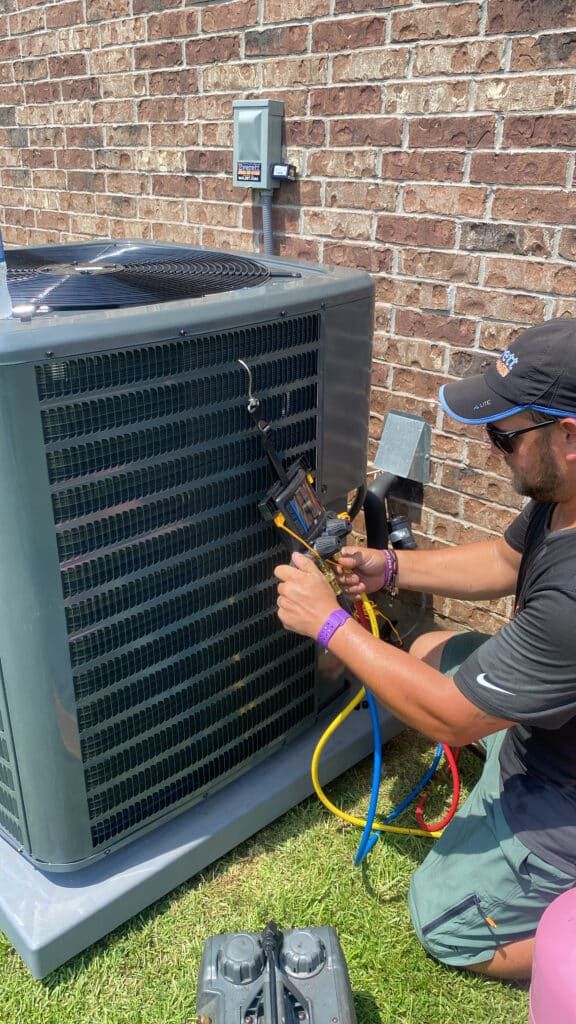 A technician in a gray shirt and hat repairs an outdoor air conditioning unit next to a brick wall.