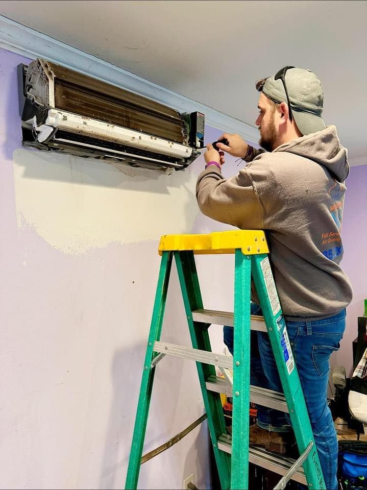 A person in a hoodie and cap stands on a green ladder, using a tool to repair or service a wall-mounted AC unit.