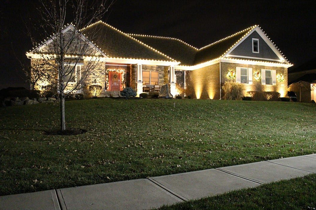 Single-story house at night outlined with white holiday lights, glowing warmly behind a dark lawn and sidewalk.