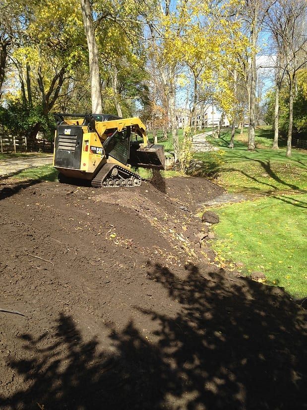 Yellow skid steer spreading dirt on a muddy path in a wooded park with trees and grass