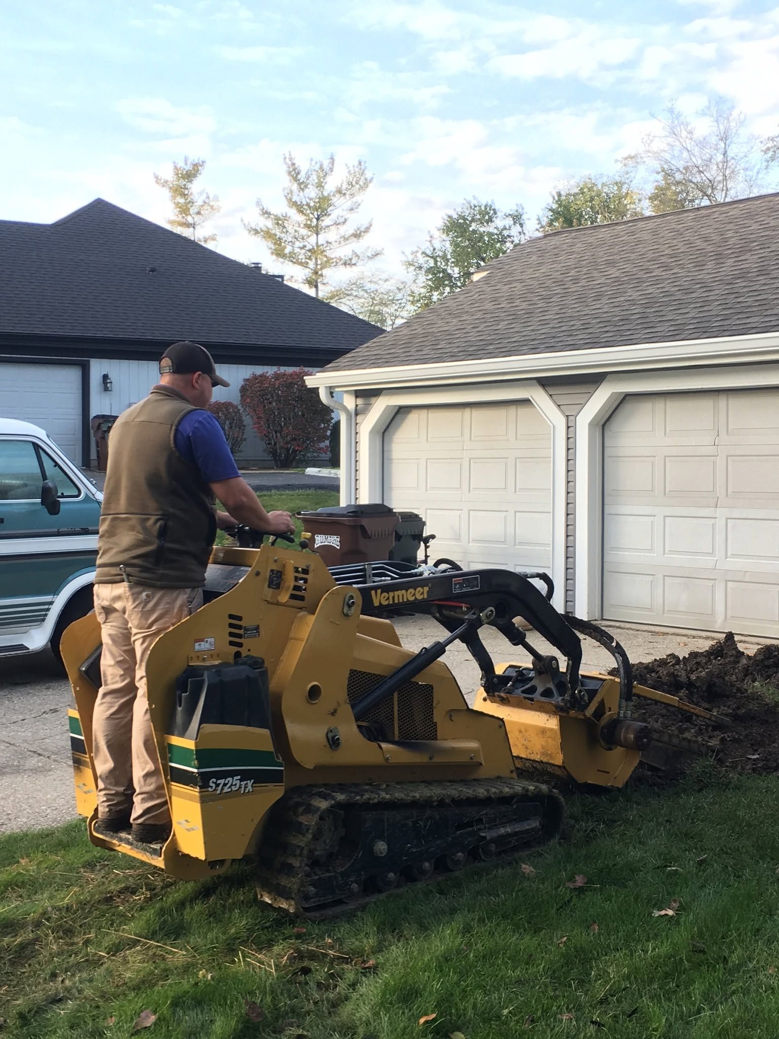 Worker standing on a yellow tracked machine trimming grass beside a white garage.