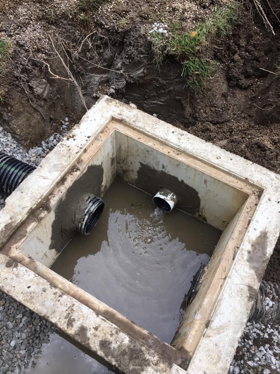 Concrete drainage box filled with muddy water beside a dirt trench and black pipe