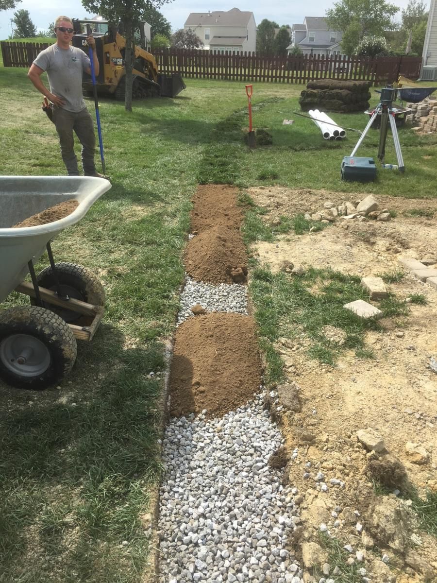 Landscaping a gravel trench beside a lawn, with workers, tools, and wheelbarrow in a backyard.