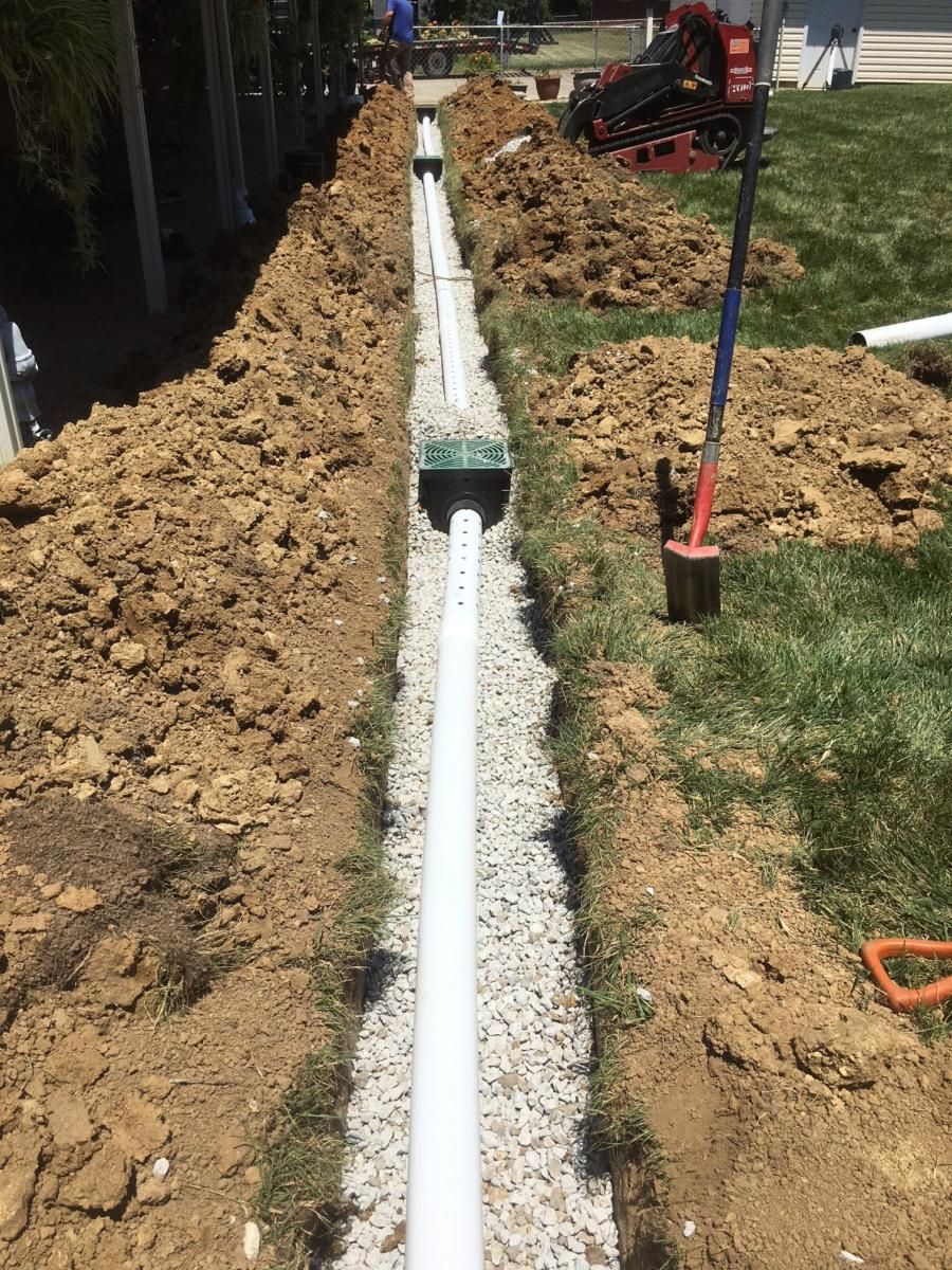 Trench with white drainage pipe and gravel, beside a grassy yard with a shovel and excavator in the background