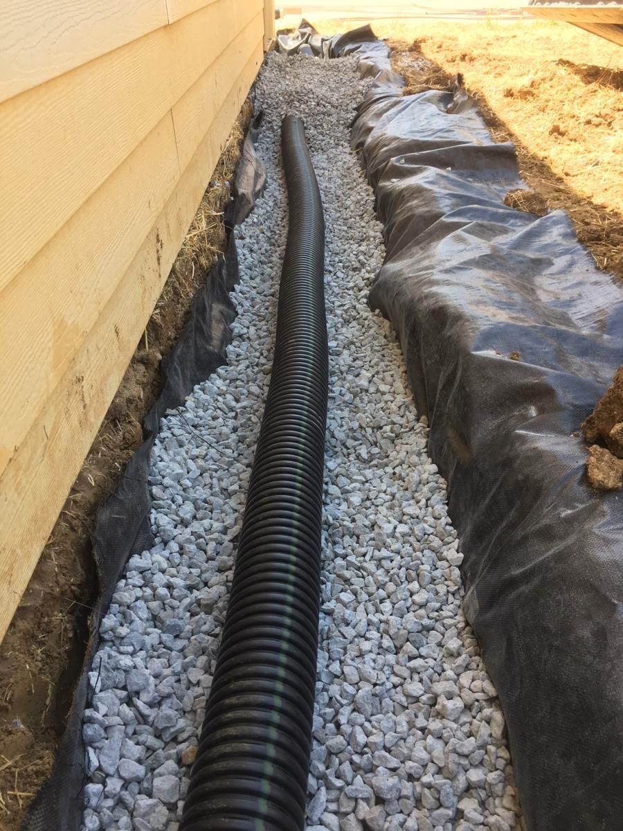 Black drainage pipe laid in gravel trench beside a building foundation, lined with black plastic sheeting.