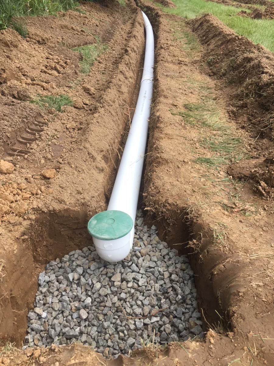 White PVC pipe in a dirt trench, ending in a green-capped drain over gravel.