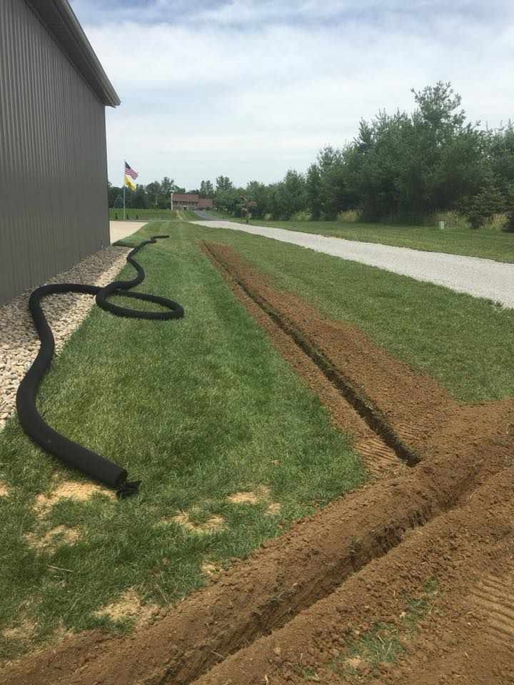 Black drainage hose beside a building, with a muddy trench cut through the grass along a roadside.