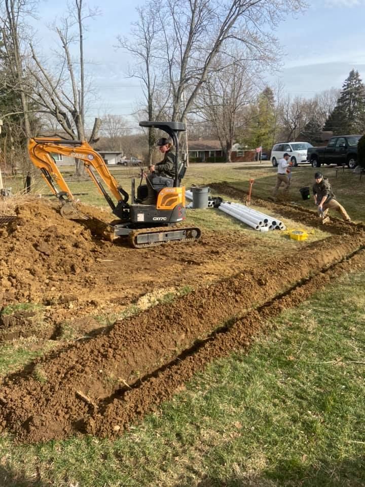 Mini excavator digging a trench in a grassy yard beside a dirt pile, with workers and a truck nearby.