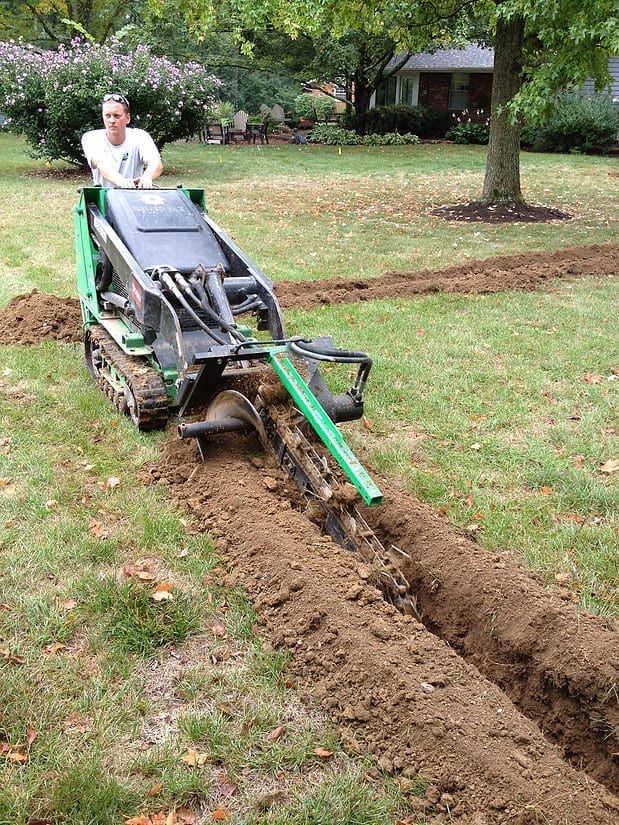 Person operating a green trenching machine digging a long narrow ditch in a grassy yard