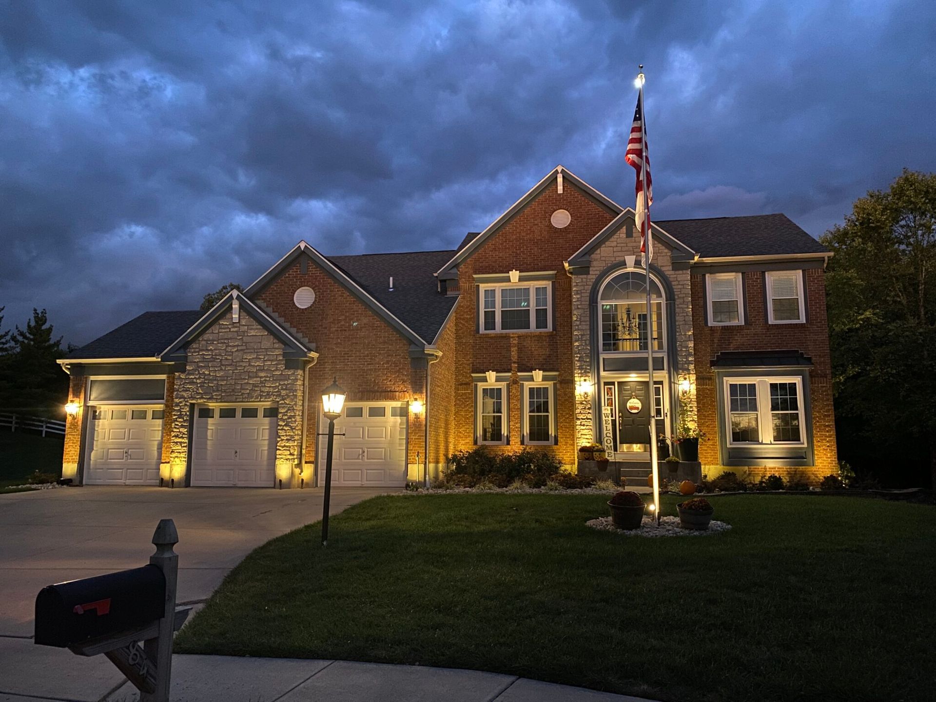 Illuminated suburban house with brick and stone exterior at dusk, U.S. flag, and driveway lights.