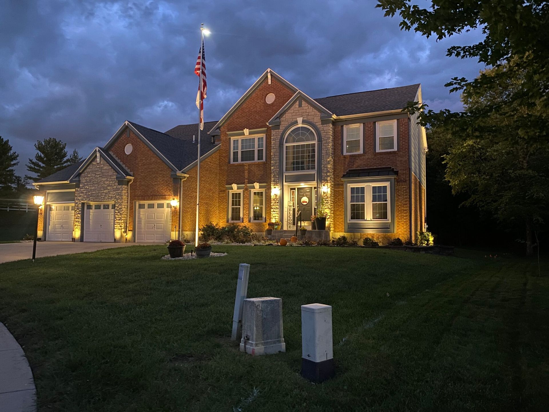 Large brick house at dusk with warm lights, an American flag, and a dark lawn in front.