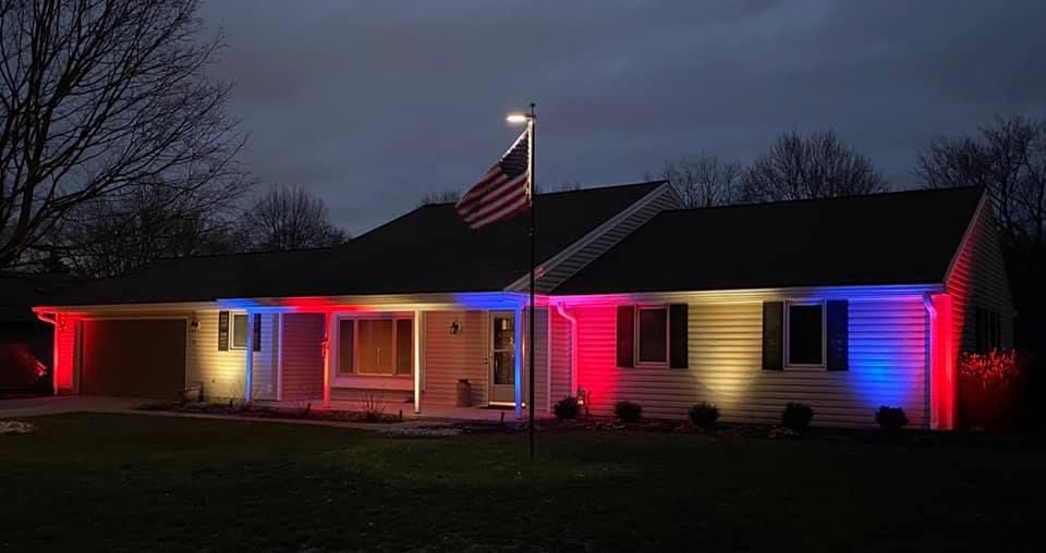 Single-story house at dusk lit with red, blue, and pink holiday lights, with an American flag flying in front.