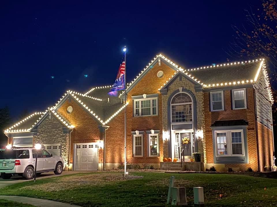 Brick house at night outlined with white holiday lights, with a lit U.S. flag and a white SUV in the driveway.
