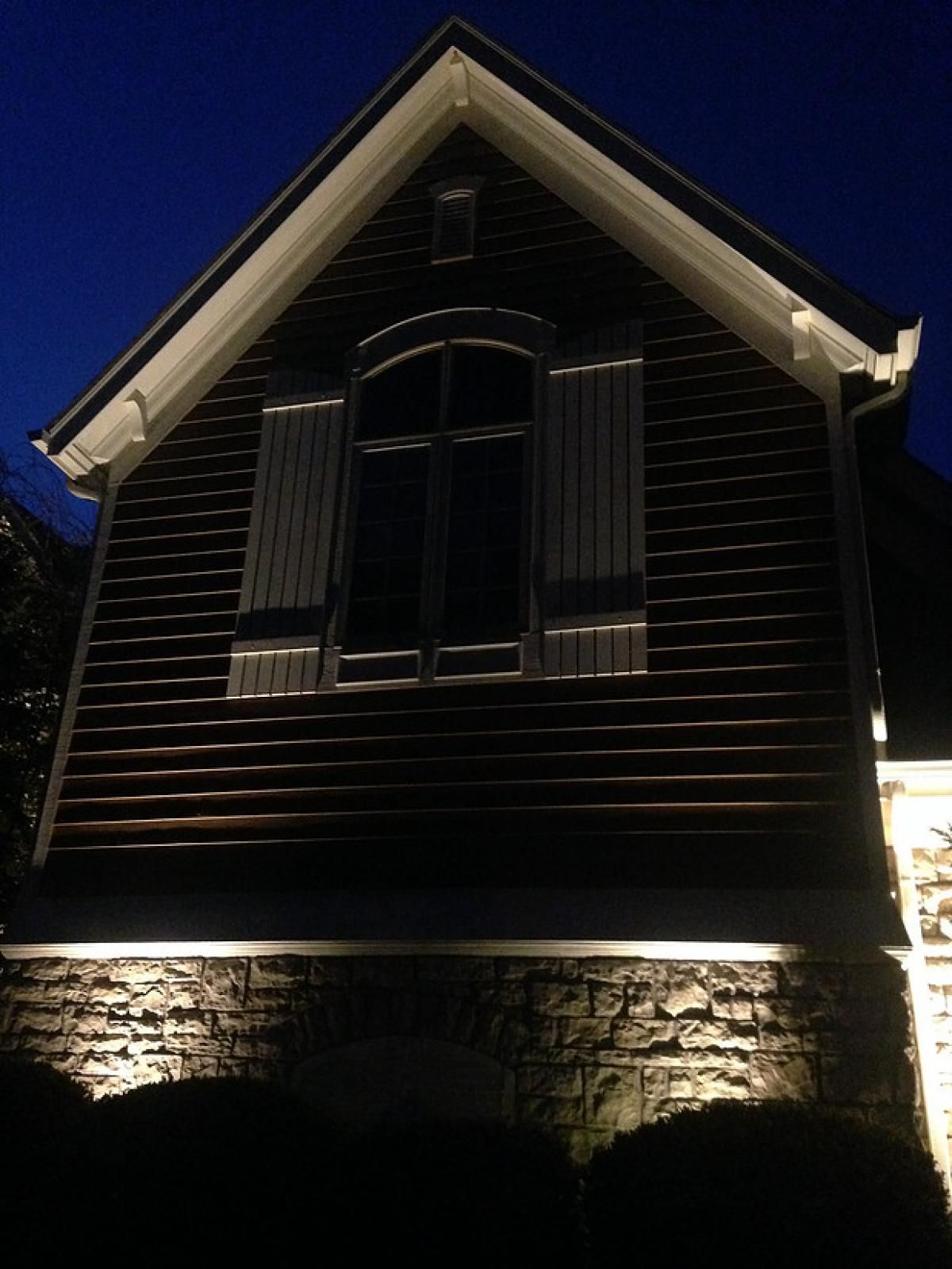 Nighttime view of a dark wooden house with lit stone foundation and a peaked roof against a blue sky