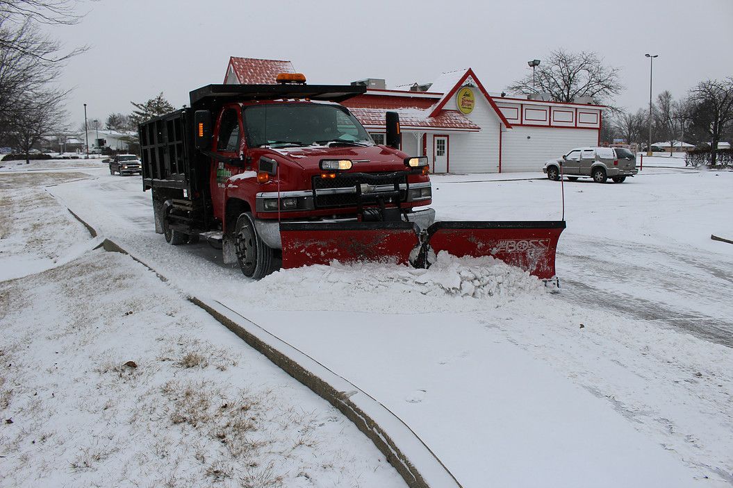 Red snowplow clearing a snow-covered road near buildings during a winter storm
