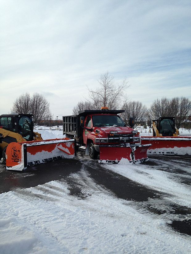 Snowplows lined up in a snowy lot, with red and orange plows facing forward under a cloudy sky.